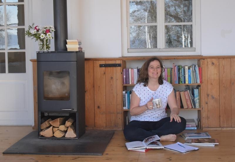 Lächelnde Frau mit Teetasse sitzt neben einem gemütlichen Ofen und vor einem Bücherregal auf dem Boden und hat Notizen um sich