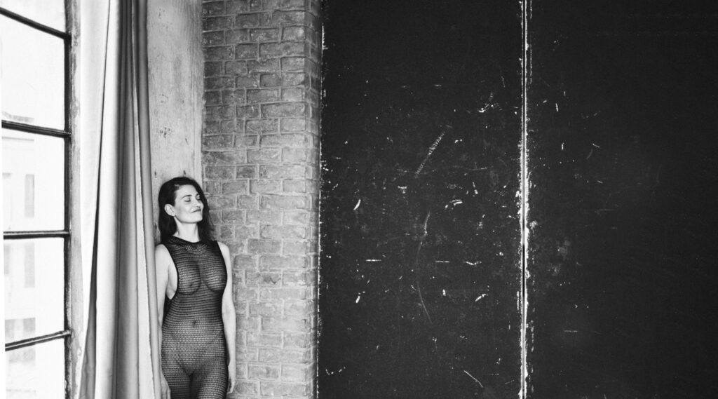 Frau in durchsichtigem Kleid lehnt an der Wand in einem loftartigen Raum